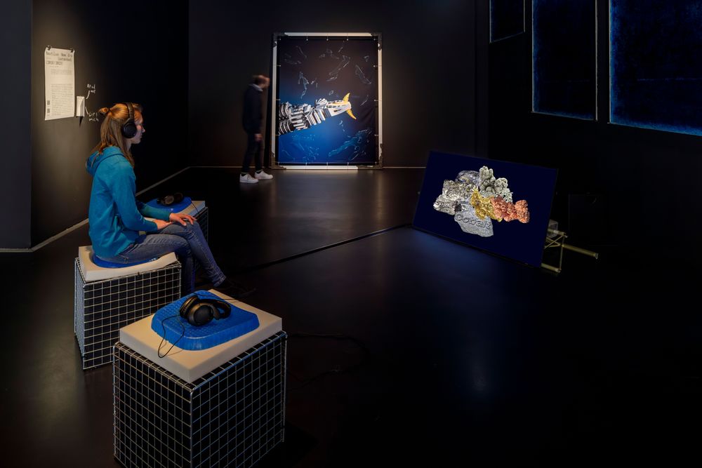 Die Ausstellungsansicht zeigt eine Frau die Kopfhörer trägt und auf einem Hocker sitzt. Sie schaut auf einen Bildschirm, auf dem ein Video abgespielt wird. 