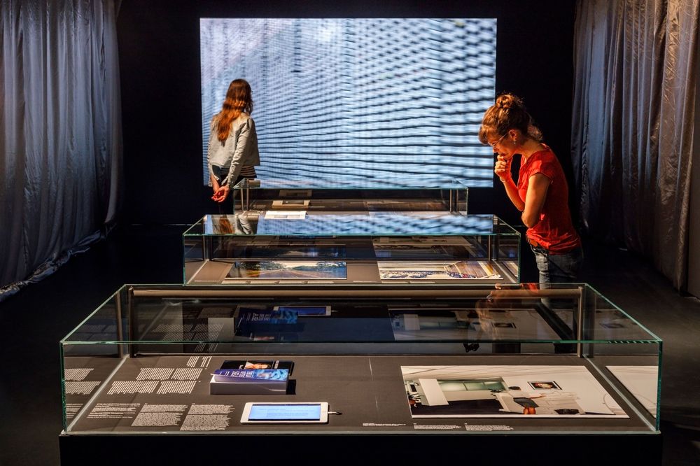 Die Ausstellungsansicht zeigt eine Frau, die vor sich ein Bild in einem Glaskasten betrachtet. Dahinter steht eine weitere Frau, die sich eine Videoarbeit anschaut.