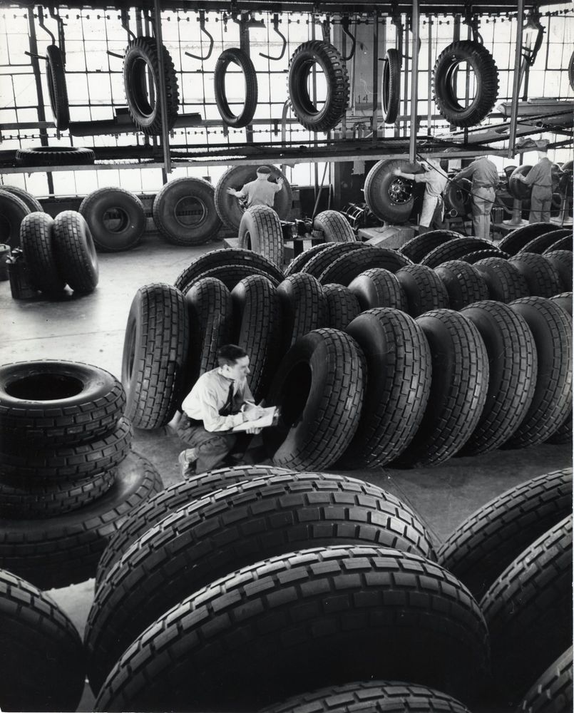 Die schwarz-weiß Fotografie zeigt eine Reifenfabrik, im ganzen Bild liegen oder Hängen Reifen an verschiedenen Stellen. Einige Reifen werden von Männern inspiziert.