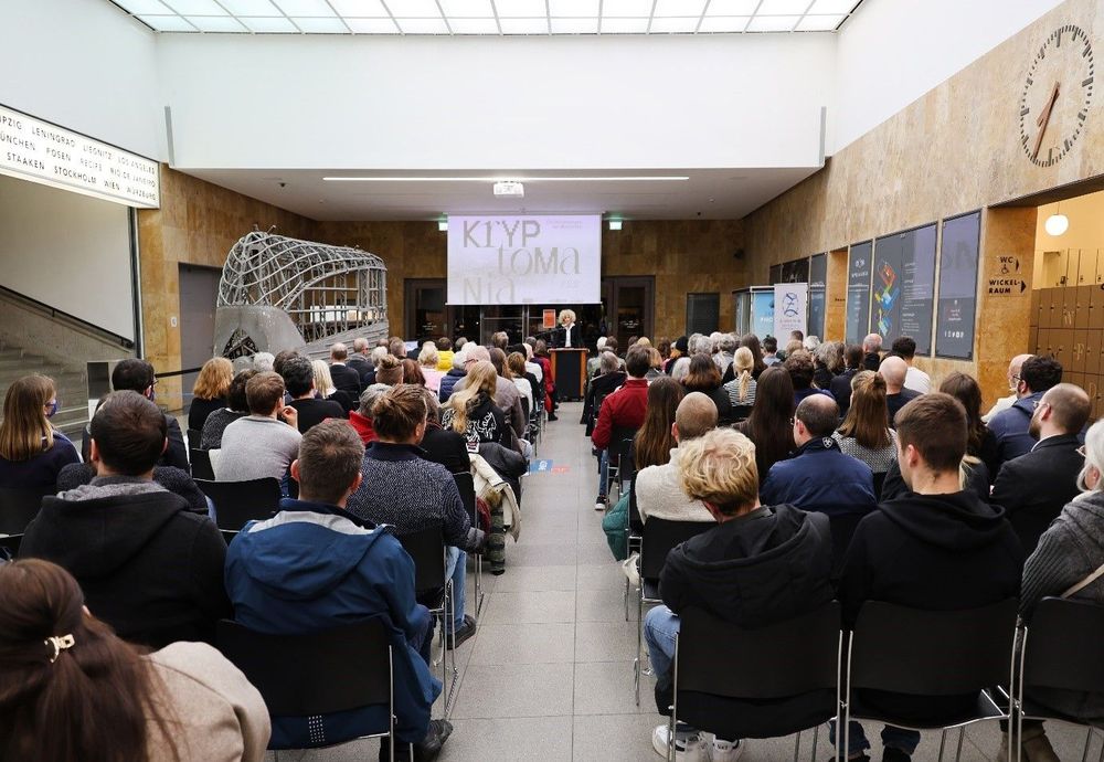 Veranstaltung in einer gut besuchten Halle mit Reihen von Zuhörer*innen, die auf eine Bühne mit Redner*innen und Präsentationsleinwand blicken.