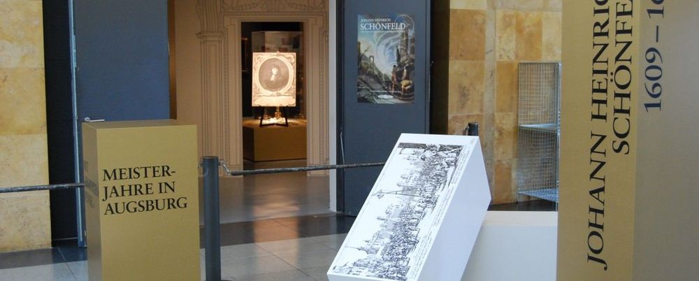 Die Aufnahme zeigt die Eingangshalle des Zeppelin Museums und den Eingang zur Ausstellung. In der Eingangshalle befinden sich verschiedene aufgestellte Blöcke, auf denen sich Schriftzüge thematisch zur Ausstellung befinden.