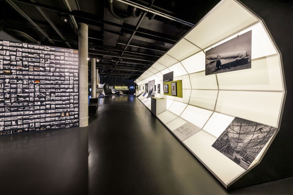 Eine dunkle Raumansicht mit einer halbrunden leuchtenden Wand, an der Fotos hängen, davor Personen