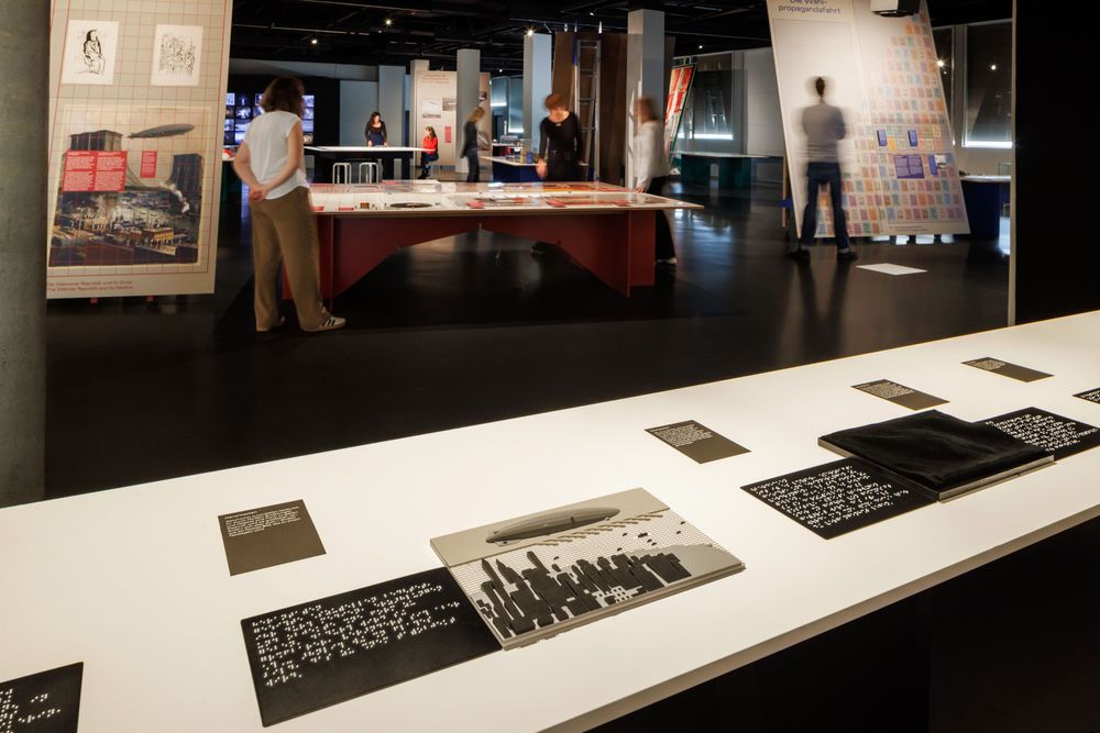 Ausstellungsansicht mit einem Tisch mit Tastmodellen im Vordergrund. Im Hintergrund sind weitere Tische mit Exponaten sichtbar, an der Rückwand leuchten mehrere Bildschirme.