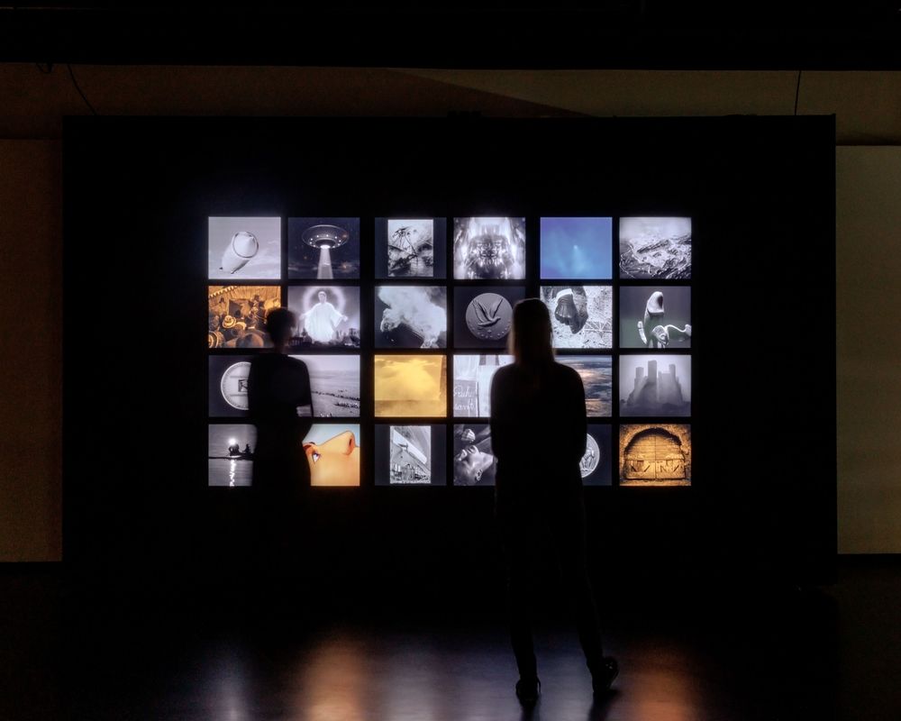 Ansicht vom Kunstwerk Cult [Zeppelin]von Jonas Englert. 24 Bildschirme mit verschiedenen Motiven vor einem schwarzen Hintergrund. Zwei Besucherinnen stehen als Silhouetten davor. 
