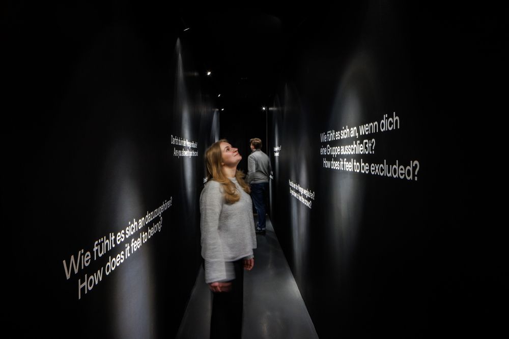 Das Foto zeigt einen dunklen Tunnel mit weiß geschriebenen Fragen an den Wänden. Mehrere Personen sind darin, im Vordergrund eine blonde Frau. Sie liest die Frage an der Wand: Wie fühlt es sich an, wenn die eine Gruppe ausschließt?