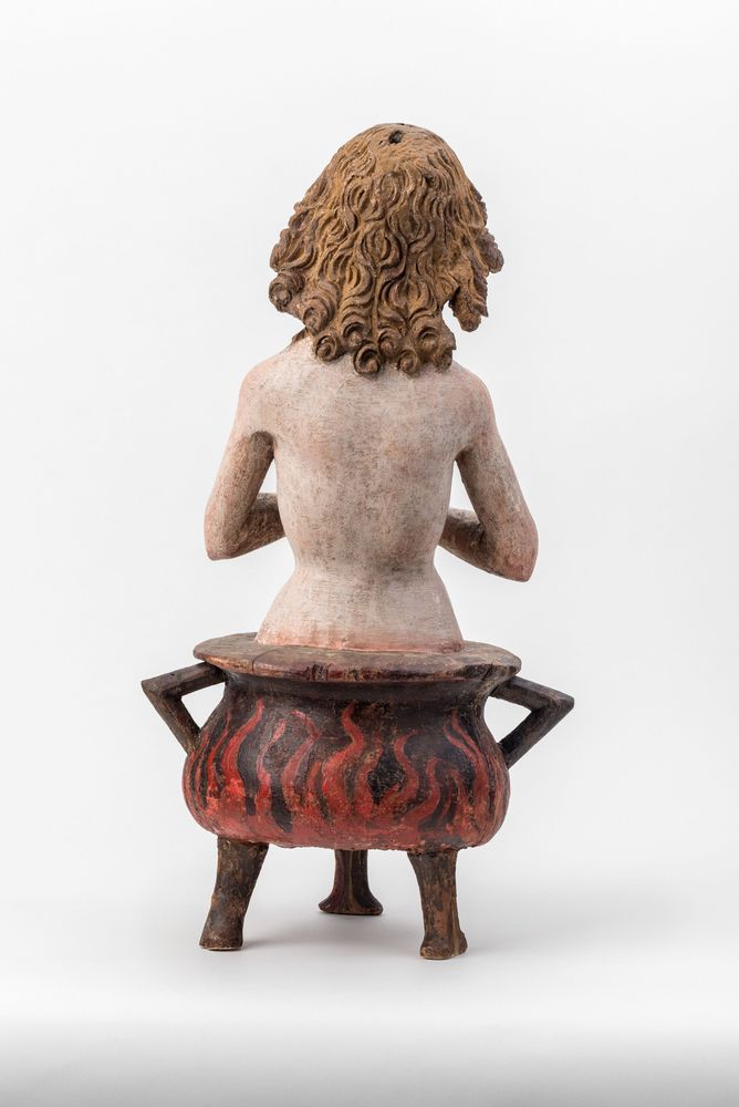 Die Aufnahme zeigt die Skulptur eines Jungen der mit betenden Händen in einem brennenden Kessel sitzt..