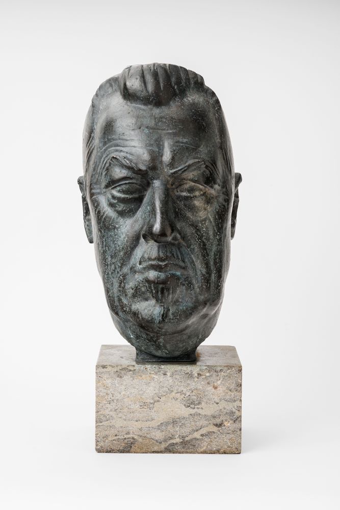 Eine Metall-Büste von Hugo Eckener. Die Büste steht auf einem viereckigen Marmorsockel, seine Höhe misst ca. 8 cm. Eckener ist im Alter von 66 Jahren dargestellt, mit schlankem Kopf, anliegenden Ohren und Geheimratsecken. Das Haar ist nach hinten gekämmt.
