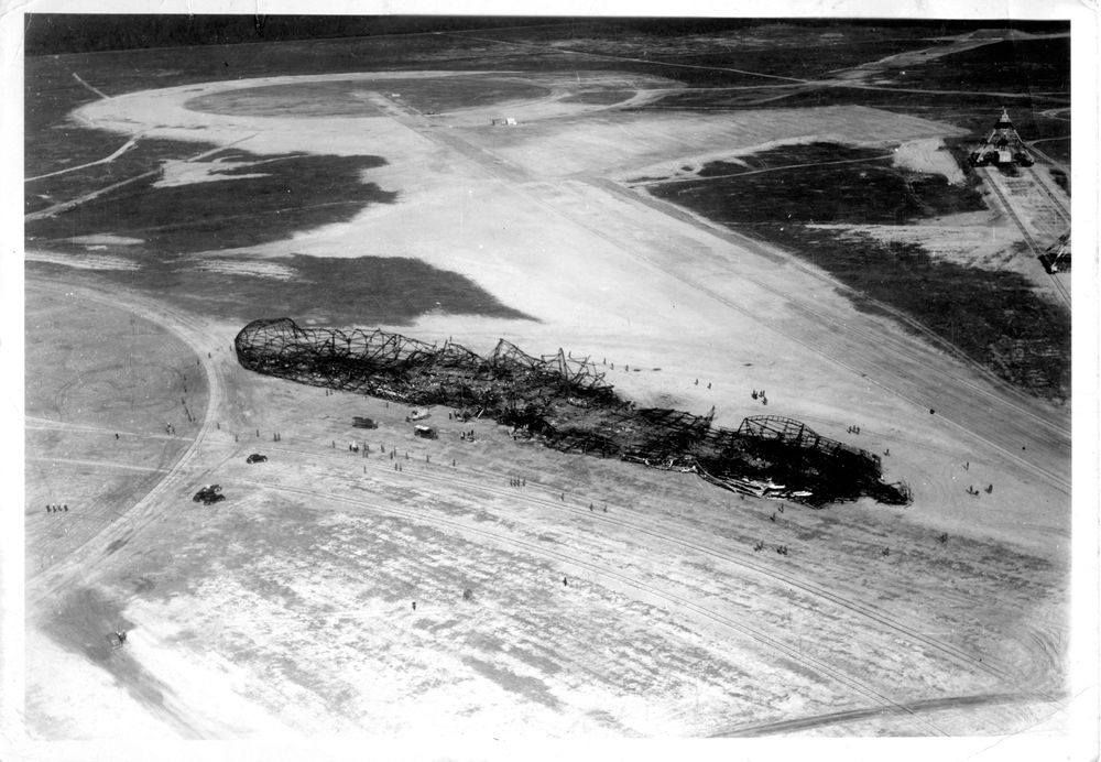 Das ausgebrannte Luftschiff von oben betrachtet, liegt auf dem Landeplatz in Lakehurst. Zahlreiche Menschen stehen drum herum. Auch einige Fahrzeuge sind zu sehen.