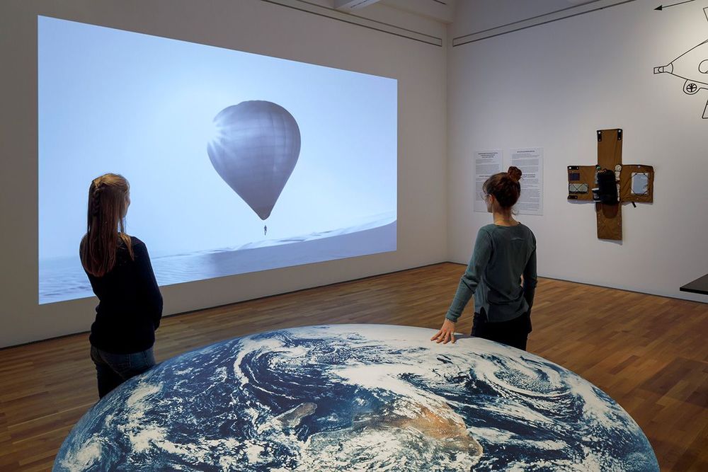 Die Ausstellungsansicht zeigt zwei Frauen, die an einem Tischplatte stehen, die wie die Erde aussieht und auf das Bild eines Heißluftballons schauen, welches an die wand projiziert ist.