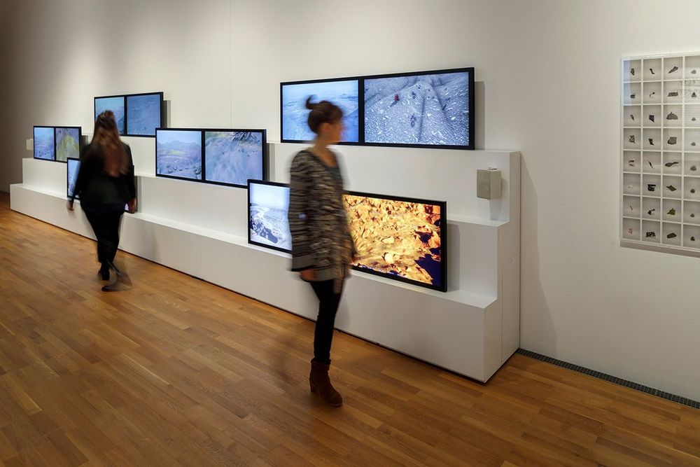 Die Ausstellungsansicht zeigt zwei Frauen die sich in gegengesetzter Richtung voneinander wegbewegen und dabei an verschiedenen Monitoren, die an der Wand befestigt sind, vorbei laufen.