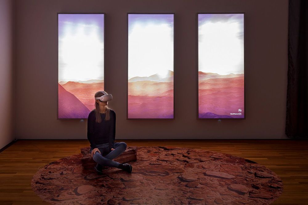 Eine Frau, die eine VR-Brille trägt sitzt auf einer Art Stein in einem Raum. Hinter ihr an der Wand befinden sich drei lange, nebeneinander hängende Bildschirme, die ein Landschaft zeigen.
