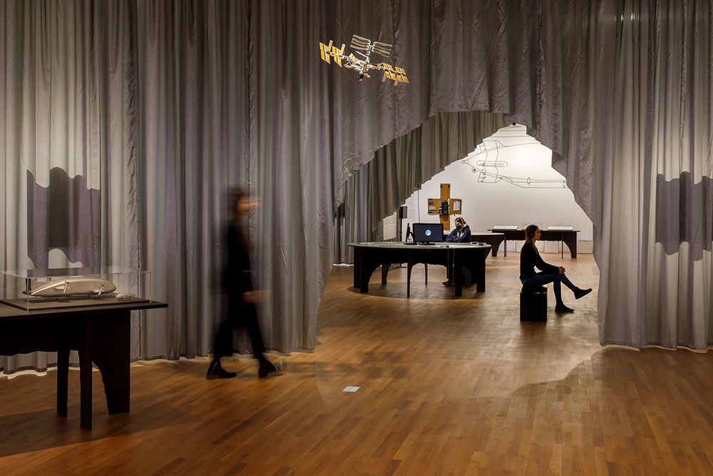 Die Ausstellungsansicht zeigt verschieden abgetrennte Bereich eines Raumes, die durch graue Vorhänge getrennt sind, welche an der Decke befestigt sind. Im hinteren Bereich der Aufnahme sitzen zwei Frauen.