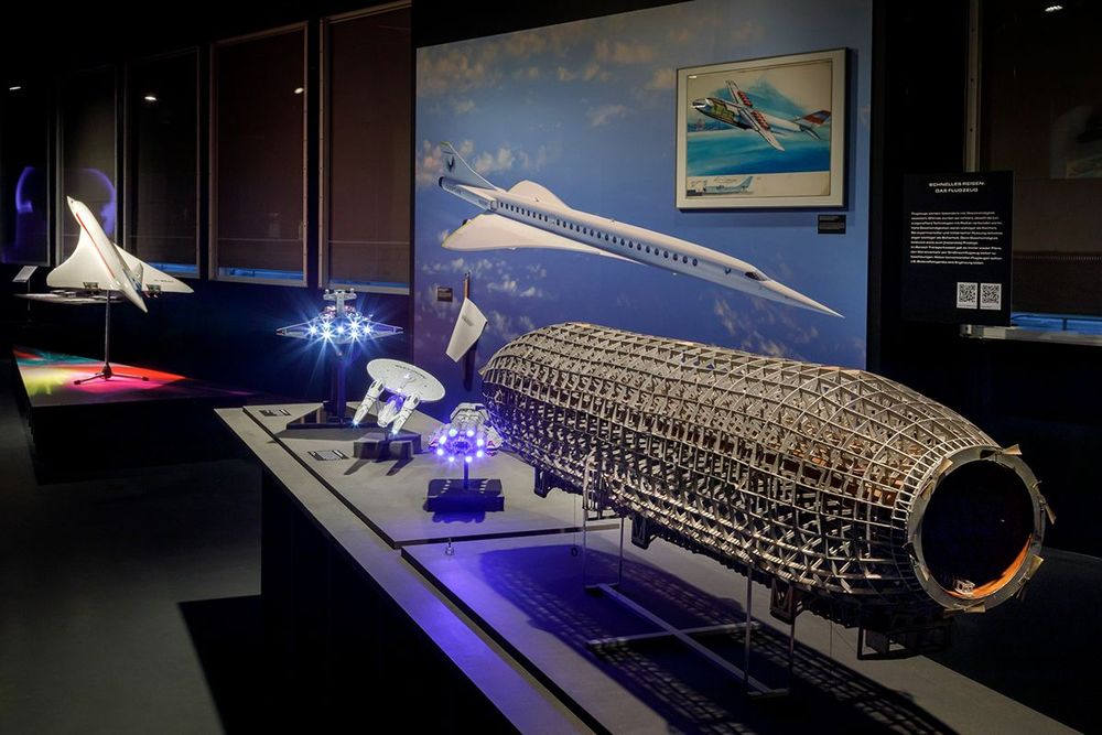 Die Ausstellungsansicht zeigt ein Podest auf dem sich verschiedene Modelle von Luftschiffen befinden. Dahinter steht ein großes Bild, das ein Flugzeug abbildet.