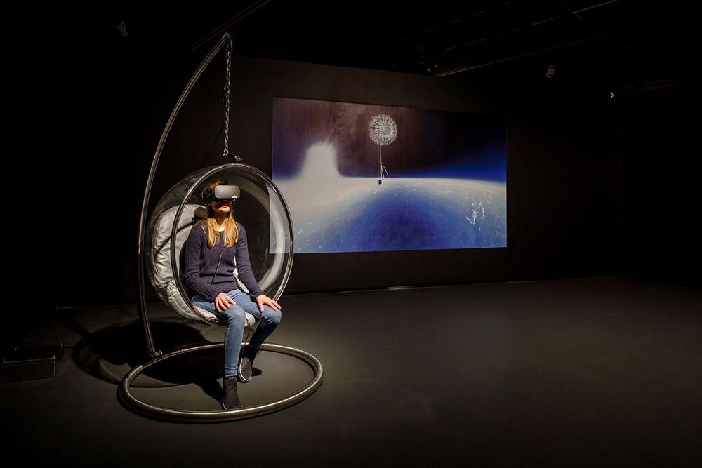 Die Ausstellungsansicht zeigt eine Frau, in einer Art Kugel-Sessel sitzt und eine VR-Brille trägt. An die Wand im Hintergrund ist ein Bild projiziert, das eine Weltraumaufnahme zeigt.