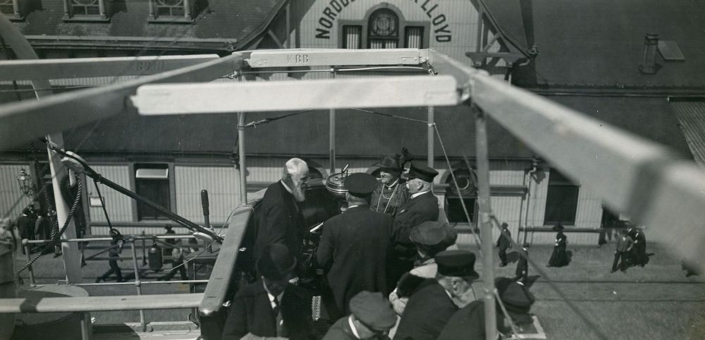 Graf Zeppelin, Prinz Heinrich von Preußen, Expeditionsteilnehmer und Gäste auf dem Dampfer „Mainz“. Im Hintergrund steht ein Gebäude mit dem Schriftzug „Norddeutscher Lloyd“.