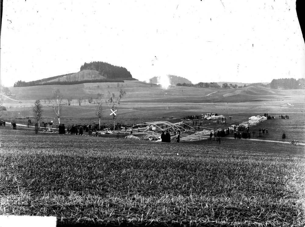 Schwarz-weiße Fotografie. Blick auf die Allgäuer Hügellandschaft. Im Bildmittelgrund ist der bereits demontierte LZ 2 zu sehen, Gerippeteile und Wrackteile sind sortiert und bereit für den Abtransport. Die gesamte Szenerie ist gesäumt von vielen Menschen.