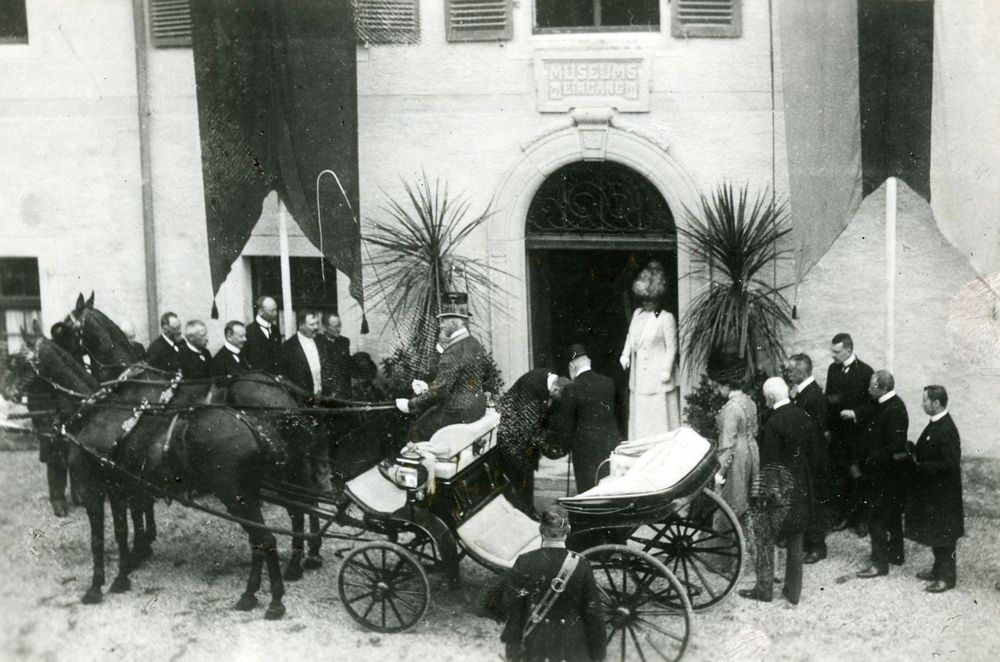 Eröffnung des Museums im Kreuzlinger Hof. Im Hintergrund der Museumseingang. Einige Menschen begrüßen das württembergische Königspaar, das in einer Kutsche angekommen ist.  