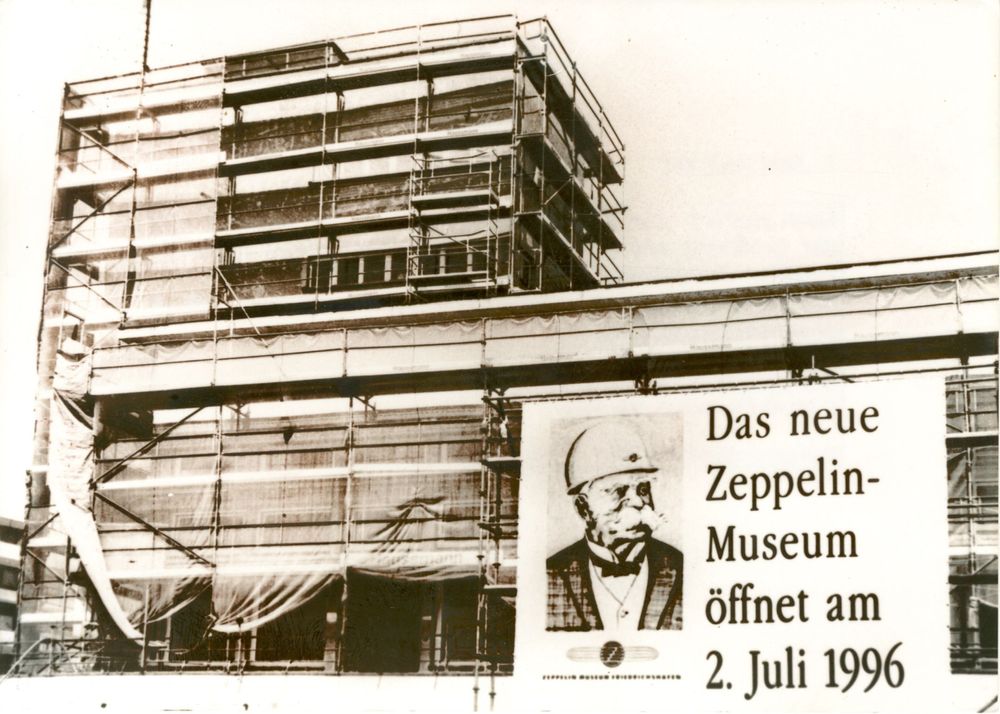 Schwarz-Weiß-Foto des Hafenbahnhofs im Umbau. Das Gebäude ist mit einem Gerüst mit Planen umstellt. Ein großes Plakat zeigt Graf Zeppelin mit einem Bauarbeiterhelm zu sehen. Rechts vom Portrait steht: Das neue Zeppelin-Museum eröffnet am 2. Juli 1996.