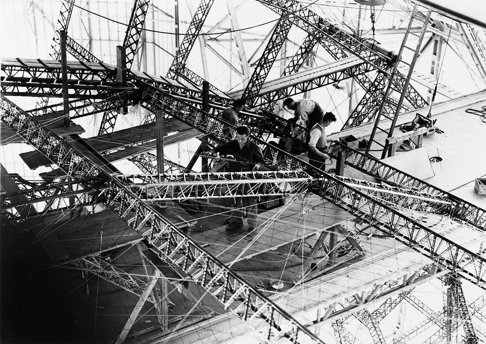 Schwarz-Weiß-Foto vom Bau des LZ 130 „Graf Zeppelin“ in der Halle. Man sieht ein Gewirr aus Streben und Drahtseilen. Auf einer Arbeitsplattform stehen vier Monteure, die an den Streben arbeiten.