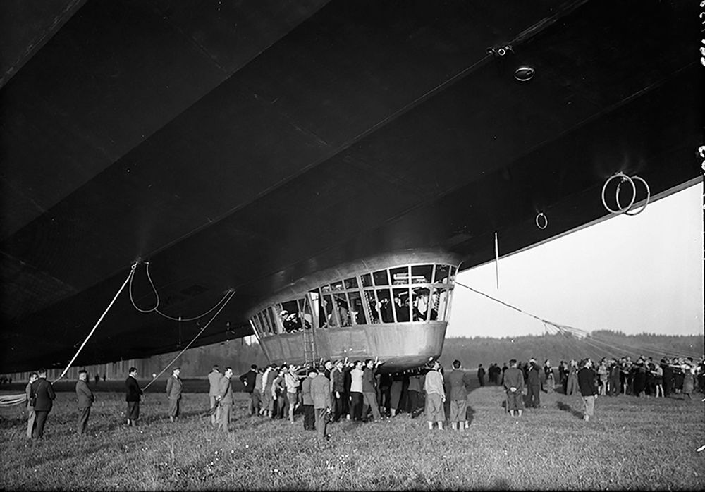 Schwarz-Weiß-Foto der Gondel des LZ 130 „Graf Zeppelin“, von der Haltemannschaft festgehalten. Mitglieder schauen aus einem der Fenster. Direkt vor ihnen ist eine Hängeleiter fixiert. Weitere Schaulustige sind ebenfalls auf dem Platz.