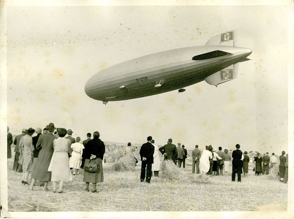 Schwarz-Weiß-Foto des LZ 130 „Graf Zeppelin“ über Viehtach im Bayrischen Wald. Unterhalb des Luftschiffs auf einem abgemähten Feld stehen zahlreiche Schaulustige zwischen einigen Heuballen.