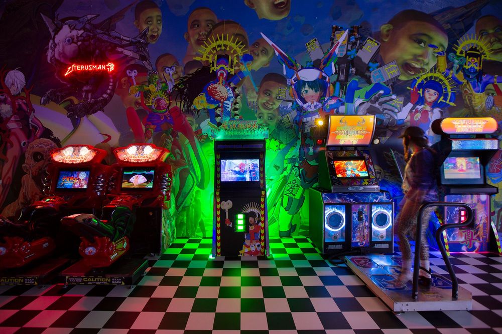 Installationsansicht mit verschiedenen Spielautomaten wie in einer Arcade. Der Boden hat ein Schachbrettmuster. 
