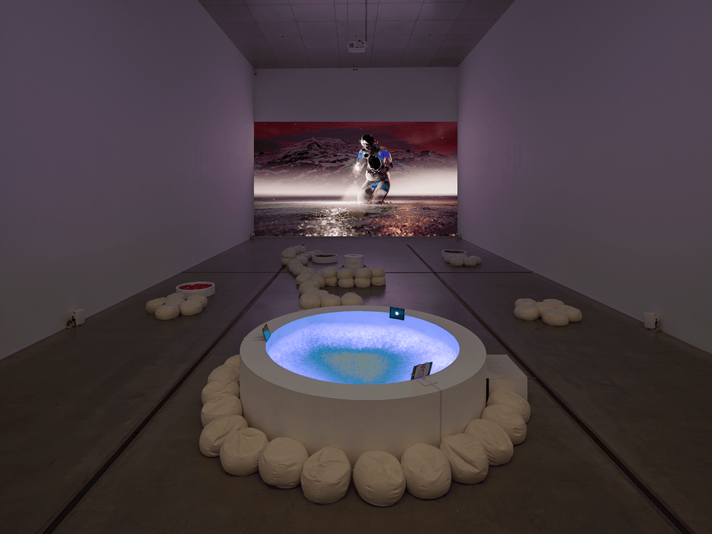 Ein runder weißer Whirlpool steht in der Mitte eines dunklen Raums mit weißen Wänden und grauem Boden. An die Rückseite des Raums wird ein Still aus der Installation projiziert. Sie zeigt eine Gestalt, die bis zu den Knien im Wasser steht.