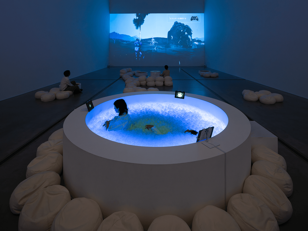 Im Vordergrund steht in weißer Whirlpool. Er ist blau beleuchtet, in ihm befinden sich weiße kleine Plastikbälle, wie in einem Bällebad. Eine Person sitzt im Whirlpool. Der Whirpool steht in einem dunklen Raum mit weißen Wänden und grauem Boden.
