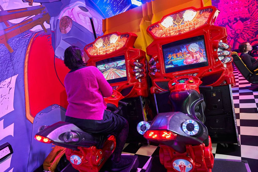 Installationsansicht auf eine Spielstation mit zwei Motorrädern. Auf dem linken Motorrad sitzt eine Frau, man sieht sie von hinten. 