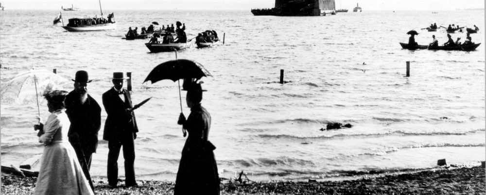 Historische Aufnahme mehrerer Personen, die am Bodenseeufer spazieren gehen. Im Hintergrund sieht man die schwimmende Halle in der Manzeller Bucht und ein paar Boote, die auf dem Wasser treiben.
