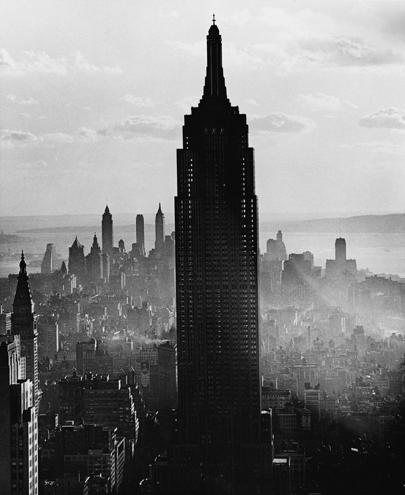 Das Empire State Building erhebt sich in der Bildmitte über die Dächer New Yorks. Es erscheint als schwarzer Schatten im Vordergrund, im Hintergrund sieht man weitere Wolkenkratzer durch Nebelschleier hindurch.