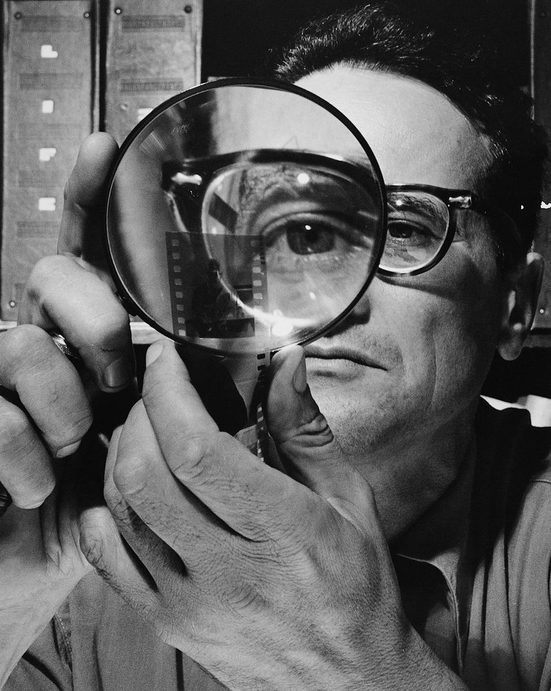 Selbstportrait von Andreas Feininger. im Vordergrund sieht man seine Hand, die eine große Lupe hält. Ein Auge wird stark vergrößert, der Rest des Gesichts ist im Hintergrund sichtbar.