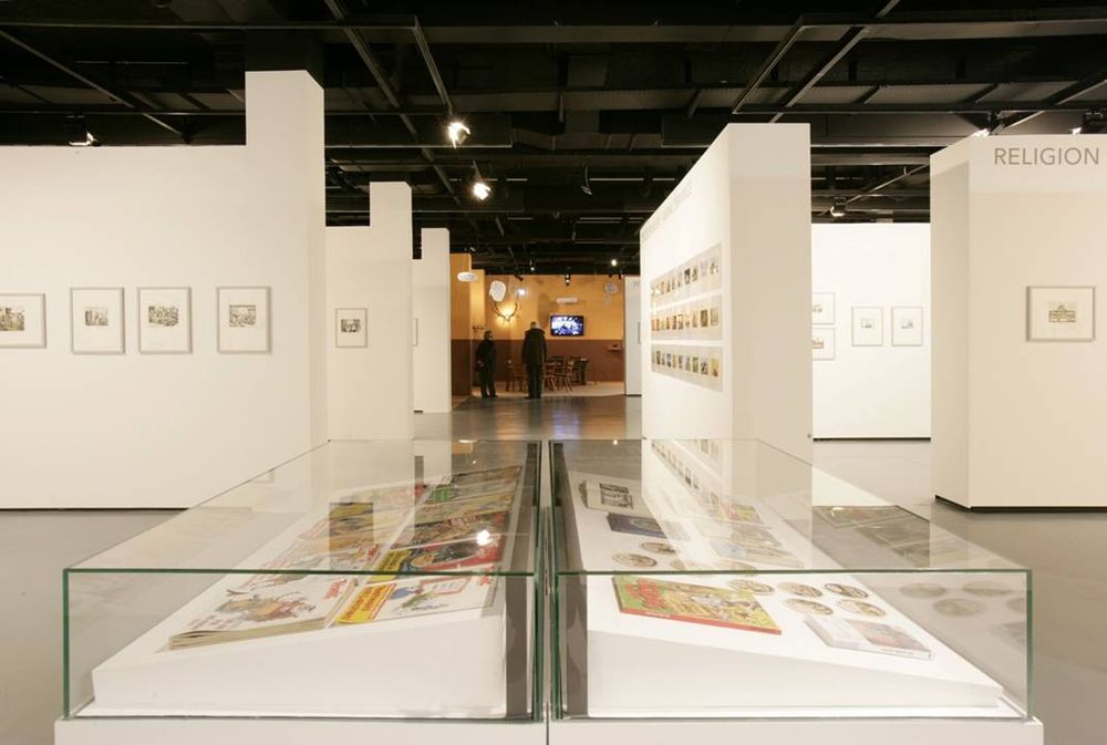 In der Mitte des Bilds stehen zwei große Vitrinen in denen sich verschiedene Exponate befinden. Dahinter sind im Ausstellungsraum weiße Wände aufgestellt, an denen verschiedene Bilder hängen. 