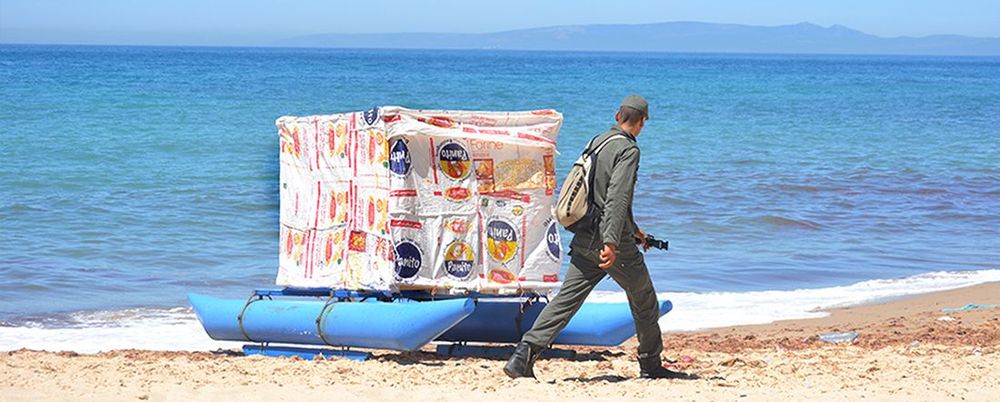 Titelbild zur Ausstellung. Ein Mann läuft am Strand an einem auf einem Boot stehenden großen Paket vorbei, das mit weiß bedruckter Folie umwickelt ist. Dahinter erstreckt sich das Meer.
