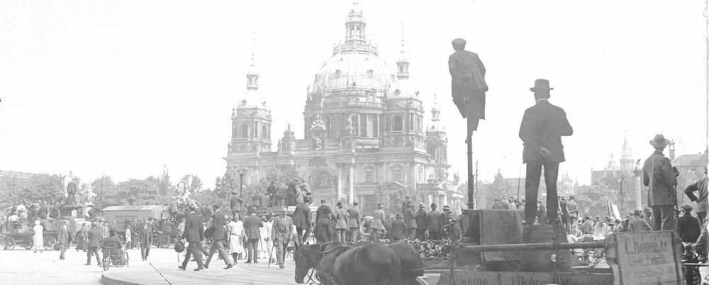 Die historische Aufnahme zeigt das Straßen-Geschehen einer Stadt. Es sind Fußgänger*inne aber auch Kutschen auf der Straße. Ein Mann ist auf eine Laterne geklettert um eine besser Sicht auf etwas in der Ferne liegende zu bekommen.