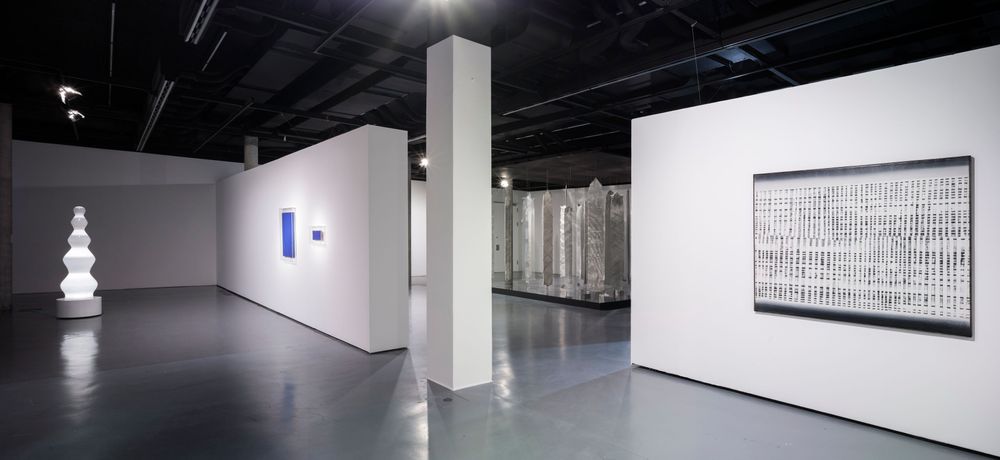 An den weißen Wänden in dem Ausstellungsraum hängen Bilder in verschiedenen Größen. Links im Bild sieht man eine leuchtende Installation, die einer großen Lampe ähnelt.