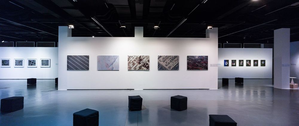 In dem Ausstellungsraum befinden sich schwarze, quadratische Hocker auf dem Boden. An der weißen Wand dahinter und an den Wänden rechts und links hängen verschiedene Bilder.