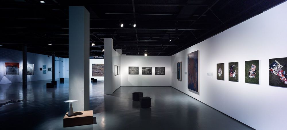 In dem Ausstellungsraum befinden sich schwarze, quadratische Hocker auf dem Boden. An der weißen Wand dahinter und an den Wänden rechts, links und hinten hängen verschiedene Bilder.