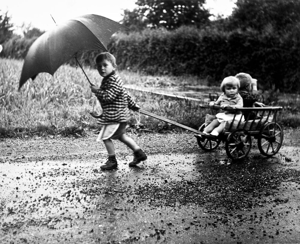 Die schwarz-weiß Fotografie zeigt drei Kinder die im Regen unterwegs sind. Das älteste Kind hält einen Regenschirm in der einen Hand und mit der anderen Hand zieht es die beiden anderen Kinder die in einem Holzwagen sitzen.