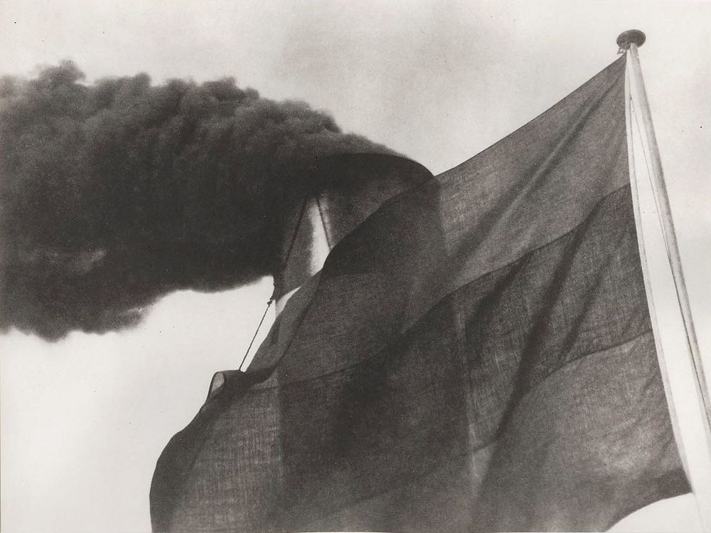 Die schwarz-weiß Fotografie zeigt die Nahaufnahme der Fahne und des Schornsteins eines Rheindampfers. Aus dem Schornstein steigt dichter Rauch auf. 