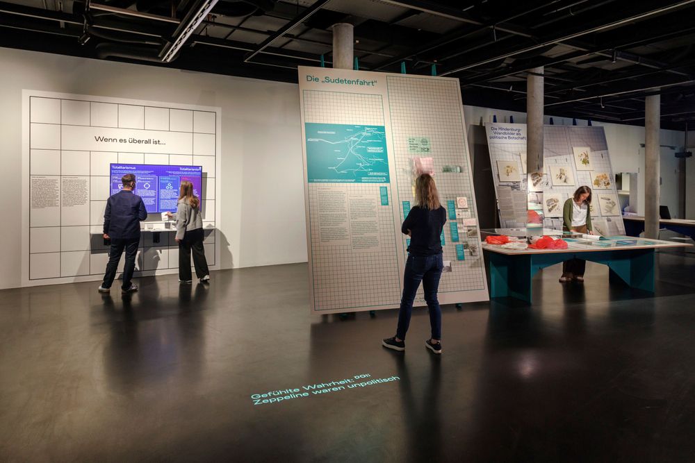 Ausstellungsansicht, mehrere Personen im Raummittelgrund, dunkler Boden, Aufsteller und Tische im Raum verteilt mit Objekten und Texten
