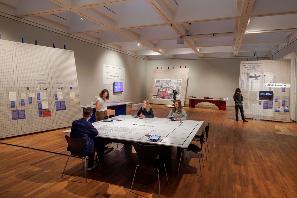 Ausstellungsansicht mit Tisch in der Bildmitte, mehrere Personen nutzen Tablet auf dem Tisch, Holzboden und helle Aufsteller an den Wänden drumherum