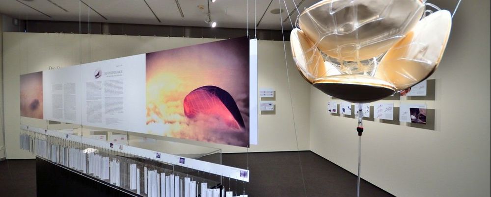 Im Vordergrund des Bildes hängt eine Installation von der Decke, links daneben im Hintergrund befindet sich eine Vitrine, über der ein Wandtafel mit Text und Bildern hängt.