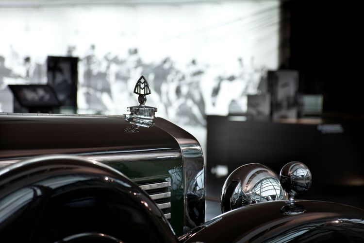 Nahaufnahme eines klassischen Maybach-Zeppelin-Automobils mit glänzendem Kühlergrill und verziertem Kühleremblem. Im Hintergrund ein unscharfes Wandbild und Vitrinen, sowie Monitore.