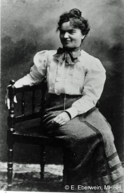 Die historische Aufnahme zeigt das Porträt einer Frau die auf einem Stuhl sitzt und leicht lächelnd links an der Kamera vorbei schaut. 