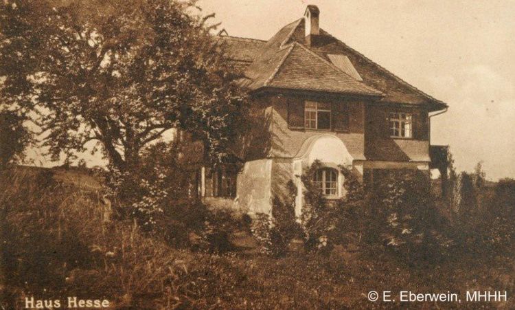 Die historische Aufnahme zeigt ein altes Haus dessen zur Kamera gewandte Wand mit Pflanzen überwachsen ist. Das Haus ist von einem Garten mit vielen großen Pflanzen umgeben. 