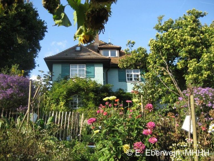 Die Fotografie zeigt ein ein hellblau gestrichenes Haus mit offenen Fensterläden. Dessen eine Wand von lauter Pflanzen bewachsen ist. Daus Haus ist umgeben von einem Garten mit sehr vielen Pflanzen und leuchtenden Blumen. 