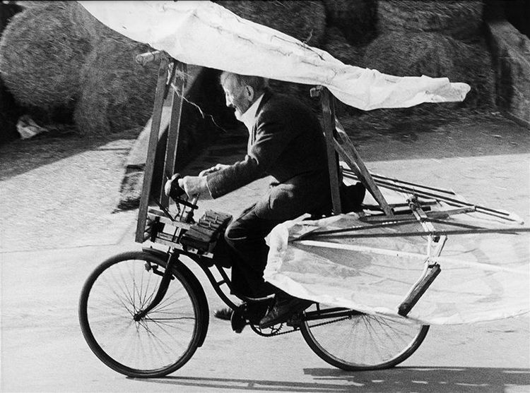 Die schwarz-weiß Fotografie zeigt einen Mann der ein Fahrrad fährt, an dem Flügel montiert sind.