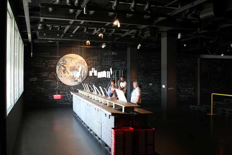 Dunkel gestalteter Ausstellungsraum mit einer langen, industriellen Theke, an der mehrere Personen Schilder ansehen. Im Hintergrund eine große, beleuchtete Darstellung des Planeten Erde an einer schwarzen Wand.
