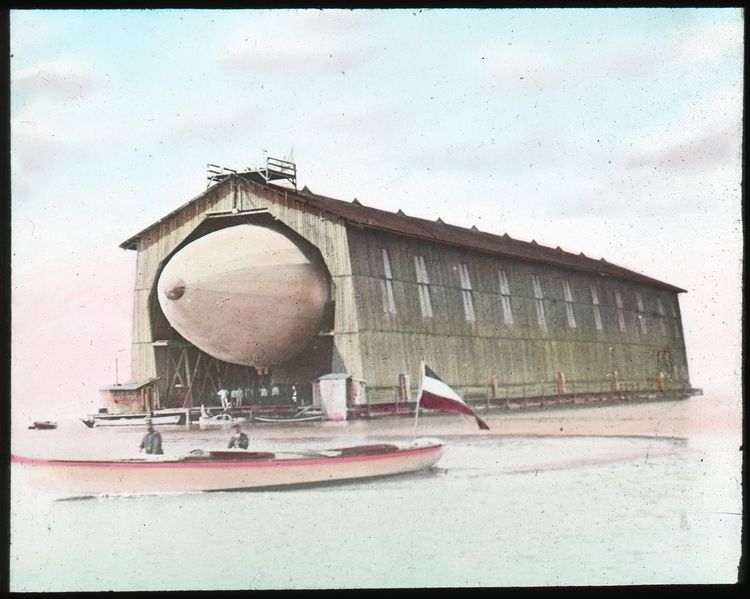 Die Bugspitze des ersten Zeppelins schaut aus der schwimmenden Pendelhalle hervor. Die Halle ist aus Holz gebaut. Im Vordergrund fährt ein rotlackiertes Boot mit einer großen Fahne vorbei.